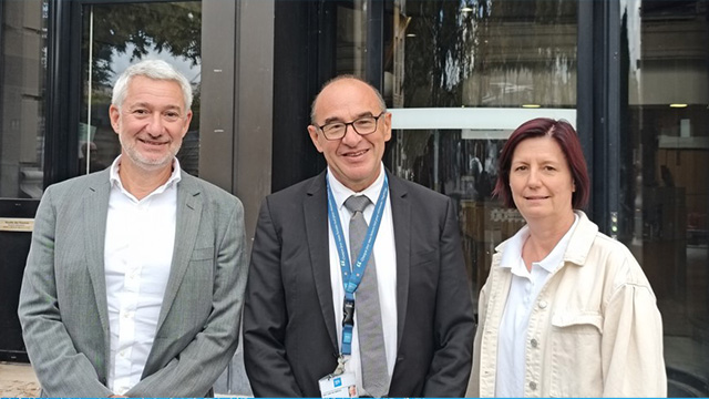 Trois personnes souriantes posent en journée devant un bâtiment aux portes vitrées. De gauche à droite : le Professeur Cotton, portant une veste gris chiné et une coupe de cheveux gris courte ; le Professeur Piriou, avec un costume bleu-gris portant un tour de cou bleu HCL ; et la Docteure Fontana, vêtue de clair avec des cheveux auburn mi-long.