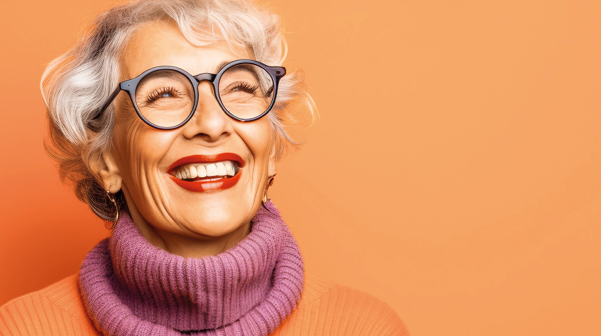 Femme souriante aux cheveux blanc portant des lunettes noirs avec un col roulé mauve sur fond orange