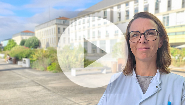 Extérieur en journée. Une professionnelle de santé de face, souriante; au niveau du buste, habillée avec une blouse blanche. À l'arrière un bâtiment de 4 étages