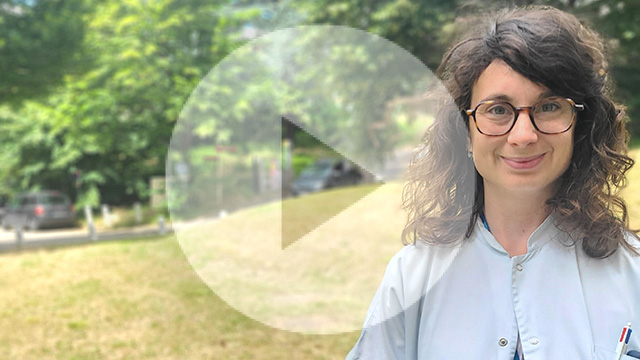 Feme brune cheveux ondulés avec lunettes et blouse blanche souriante. En arrière plan, on aperçoit une voiture de l'herbe et des arbres