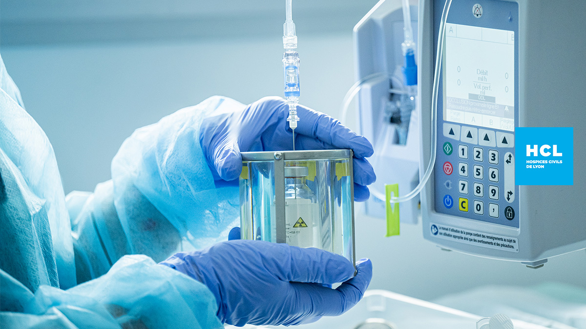 Des mains gantées de bleu tiennent un flacon contenu un liquide transparent. un fil relie le flacon à une petite machine avec un écran et un clavier