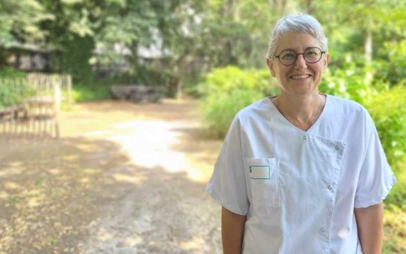 Eléonore Brocard sourit, elle a les cheveux blancs et courts, des lunettes et pose avec une blouse blanche d'infiirmière. 