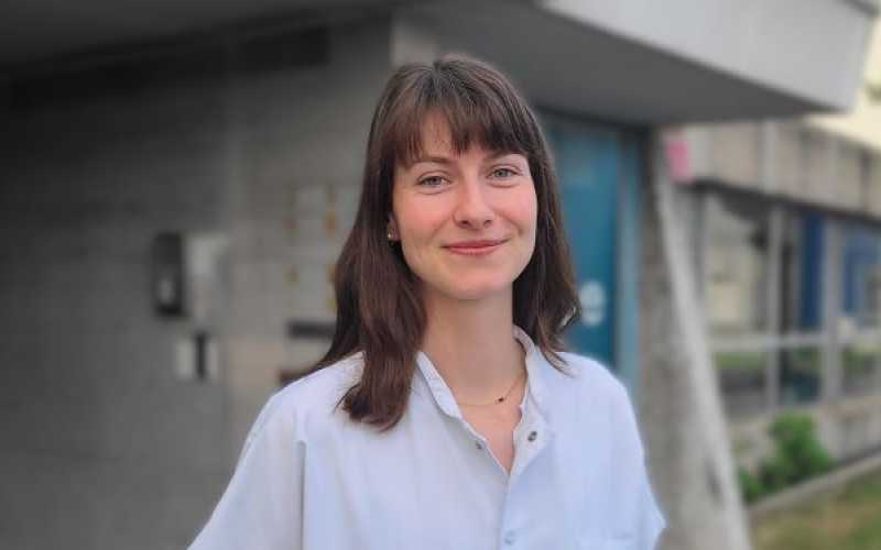 Jeune femme en blouse blanche souriante aux cheveux chatains devant un bâtiment hospitalier
