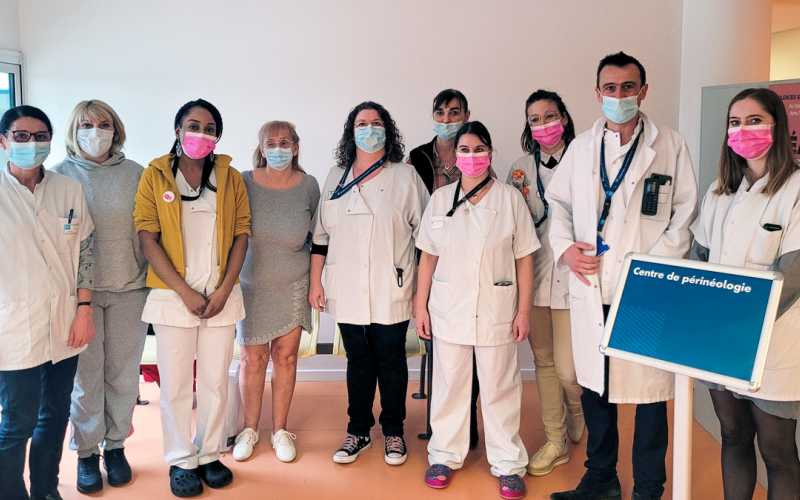 Intérieur, à l'hôpital. 10 personnes debout. 7 personnes portent une tenue médicale blanche et des tours de cou avec le bleu HCL. Tout le monde porte un masque, soit blanc, soit bleu, soit rose. Sur la droite un panneau d'affichage rectangle sur pied avec l'inscription "Centre de périnéologie"