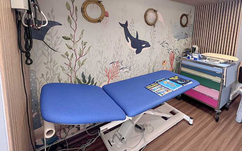 Lit de consultation hospitalier bleu avec un décor sous-marin avec des orques sur le mur du fond et des hublots