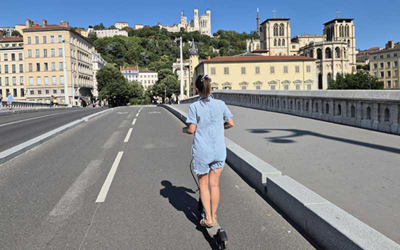 jeune fille sur une trottinette électrique sans casque de dos sur une piste cyclabe avec bâtiments et Fourvière en arrière plan