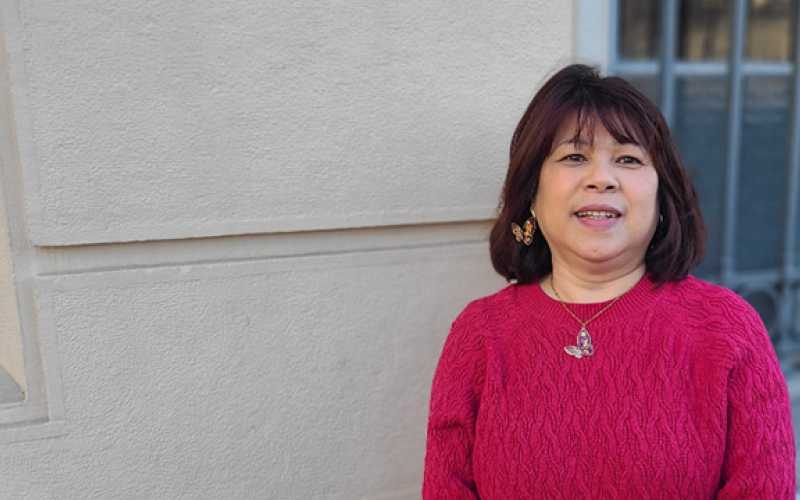 Une femme brune avec un pull rose foncé pose devant un mur et une fenêtre