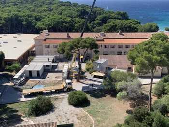vue aérienne de batiments bordés d'une foret et d'une mer. une grue évacue des algeco