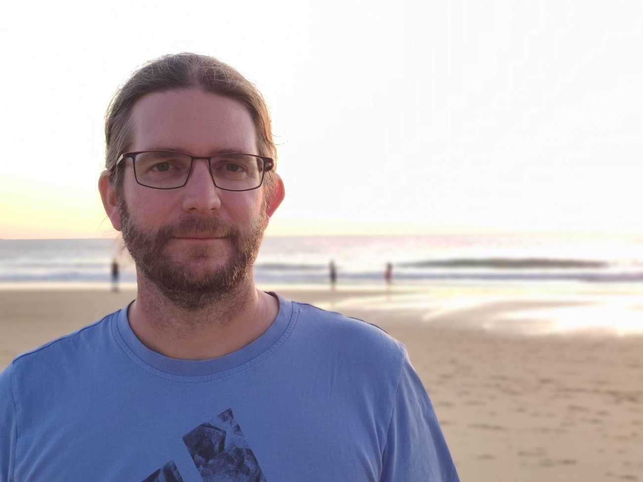 Un homme avec des lunettes, une barbe et des cheveux longs attachés, Fabien Bizot, pose sur une plage, avec la mer en fond.