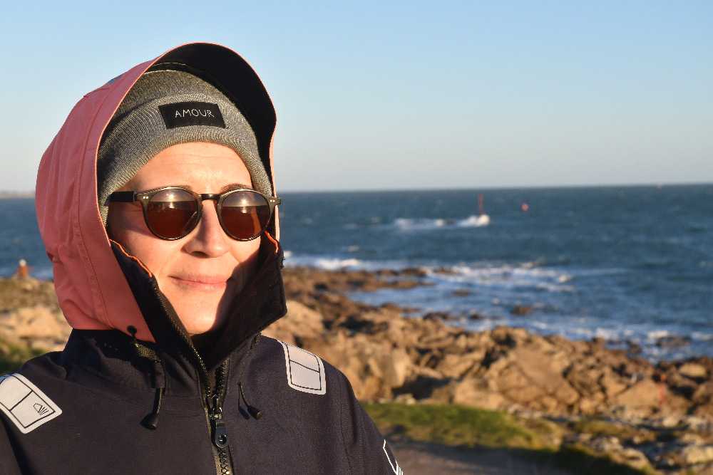 une femme avec des lunettes de soleil, un bonnet une capuche, est au bord de la mer, elle sourit