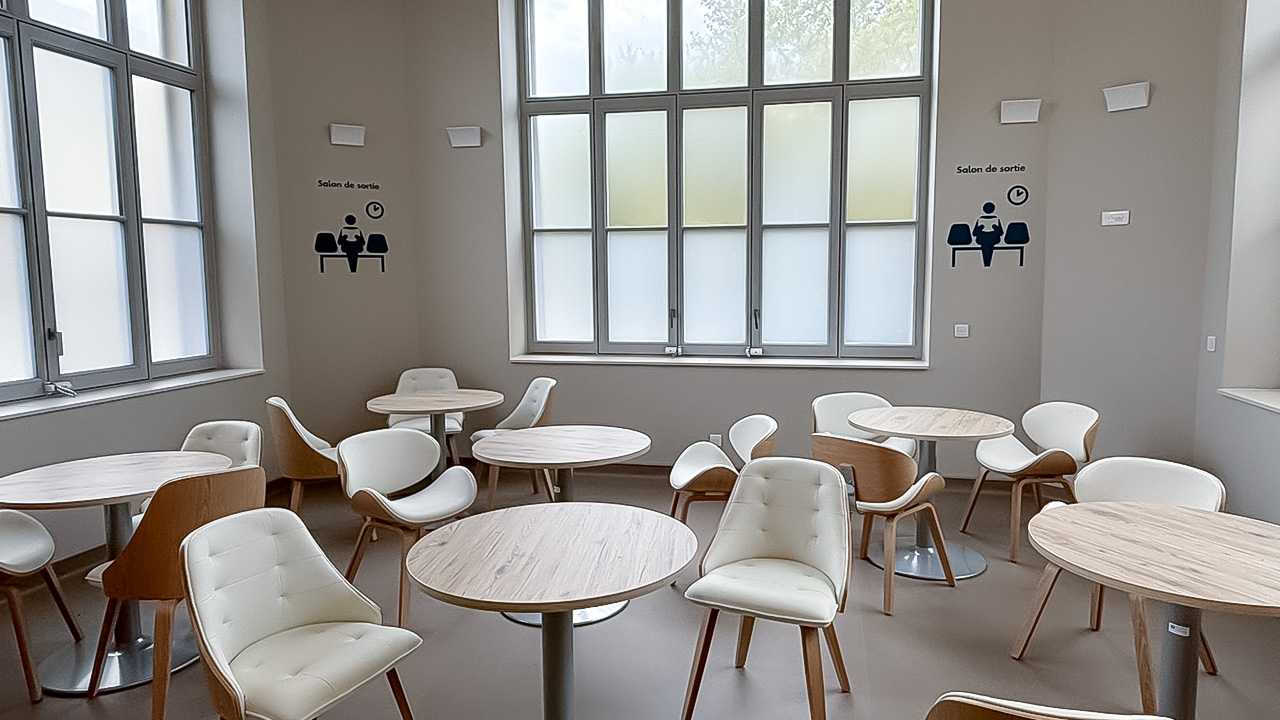 intérieur lumineux avec des grandes baies vitrées. Des tables rondes et des chaises beiges. 