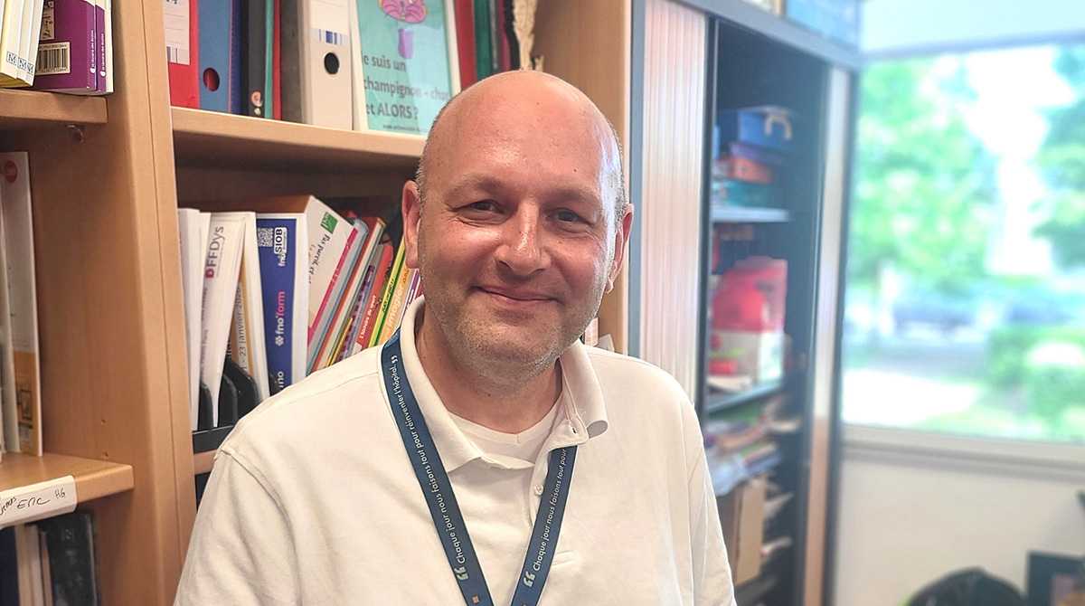 Homme souriant avec polo blanc avec en arrière plan une bibliothèque