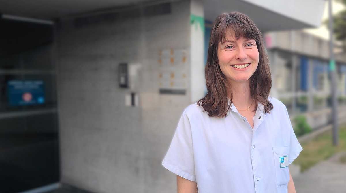 Jeune femme aux cheveux mi-longs chatains en blouse blanche devant une entrée d'hôpital