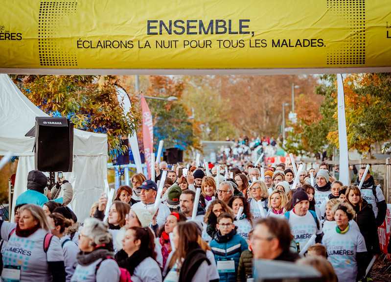 une foule de coureurs habillés de t-shirts blanc prennent le départ d'une course dans une ambiance conviviale. En fond, des arbres aux couleurs de l'automne.
