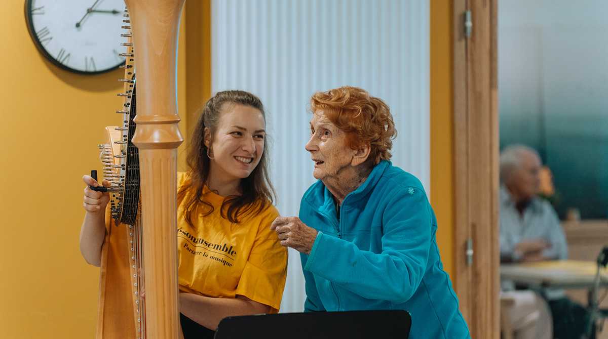 Musicienne de l'association Résonansemble avec un t-shirt jaune s’apprêtant à jouer de la harpe pour des patients à l'hôpital avec une patiente avec un haut bleu