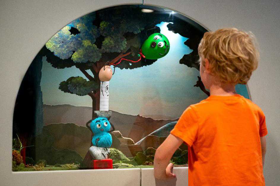 Enfant avec un t-shirt orange de dos et en face de lui un arbre avec un ballon et une peluche bleu