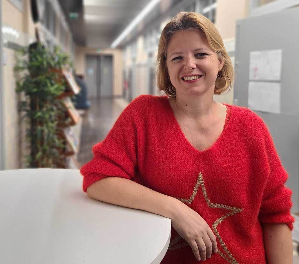 Femme souriante, aux cheveux blonds, avec un pull rouge avec une etoile. Elle est accoudée à une table dans le couloir d'un hopital.