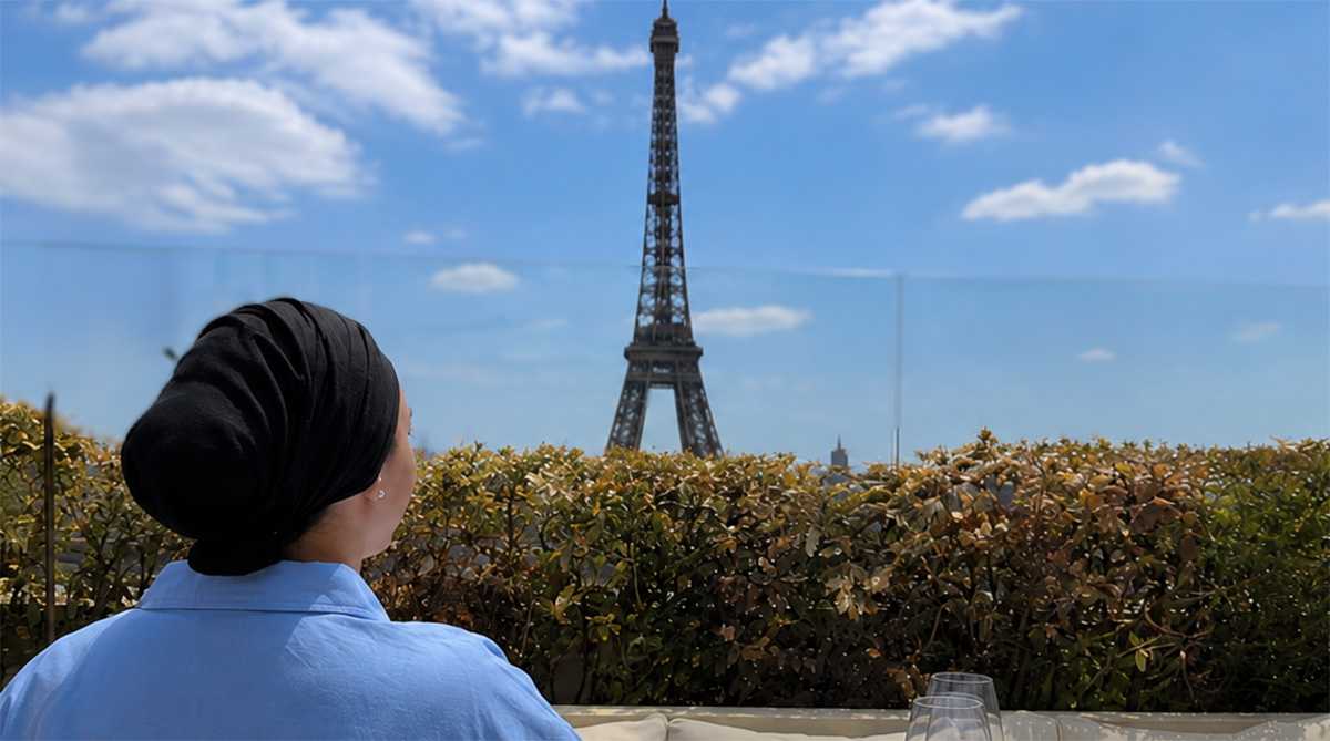 Femme de dos en blouse bleue avec un foulard noir sur la tête regardant au loin la tour Eiffel