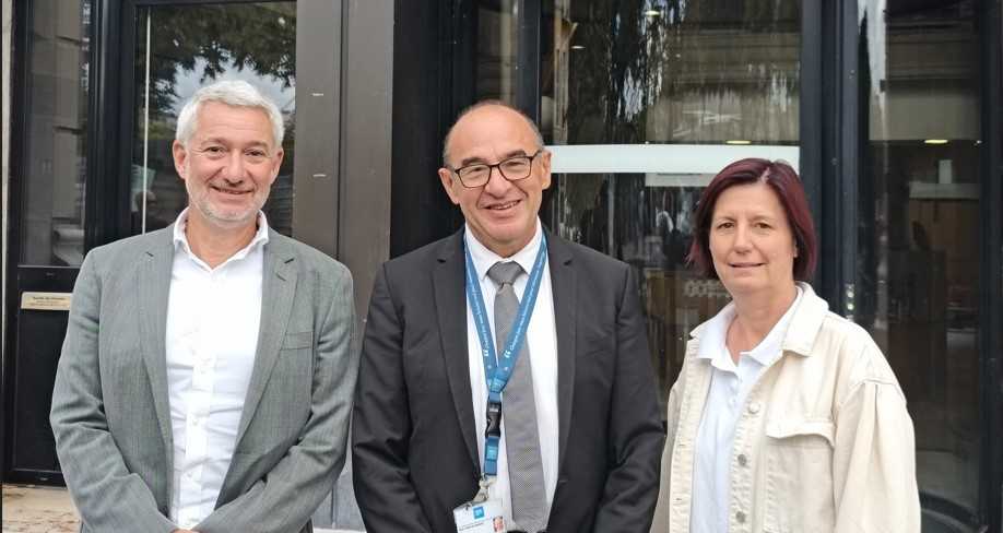 Trois personnes souriantes posent en journée devant un bâtiment aux portes vitrées. De gauche à droite : le Professeur Cotton, portant une veste gris chiné et une coupe de cheveux gris courte ; le Professeur Piriou, avec un costume bleu-gris portant un tour de cou bleu HCL ; et la Docteure Fontana, vêtue de clair avec des cheveux auburn mi-long.