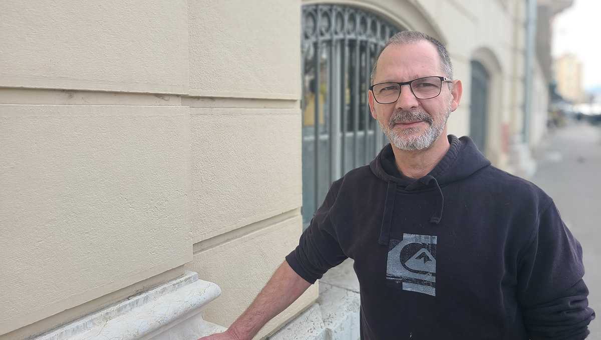 Homme avec sweet noir, barbe courte et lunettes avec une main sur un bâtiment