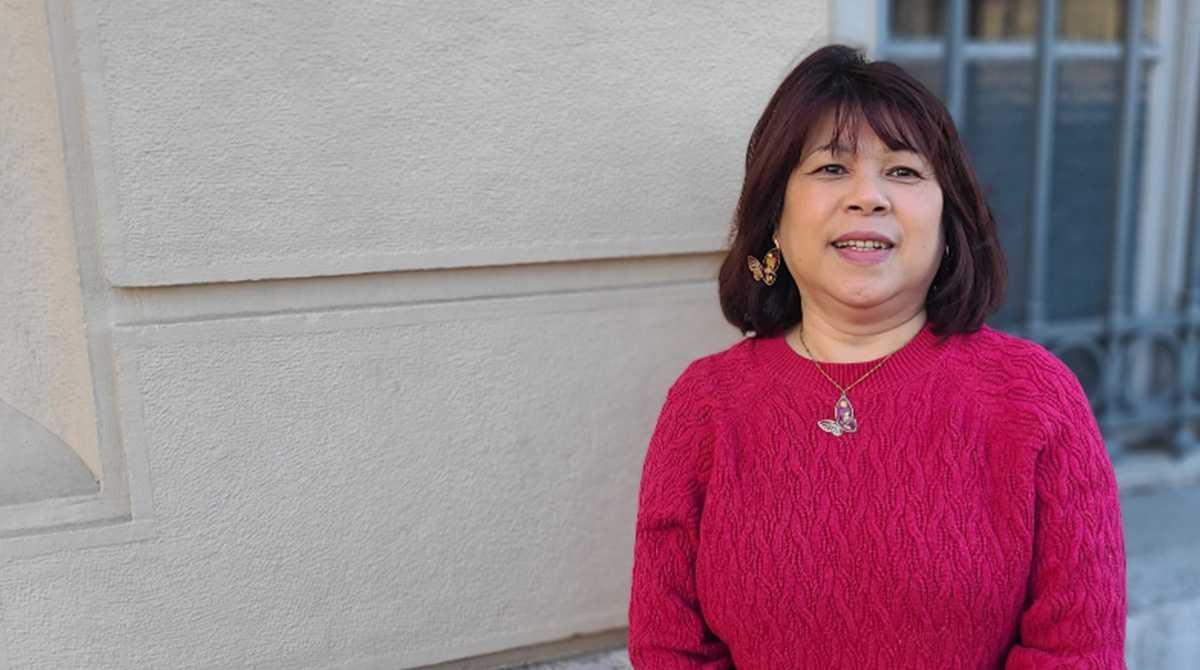 une femme brune et souriante, habillée d'un pull rose fuchsia, pose à l'extérieur, devant un mur et une fenêtre.