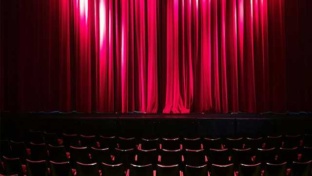 Une salle de théâtre, les sièges sont vides, le rideau rouge est fermé et éclairé.