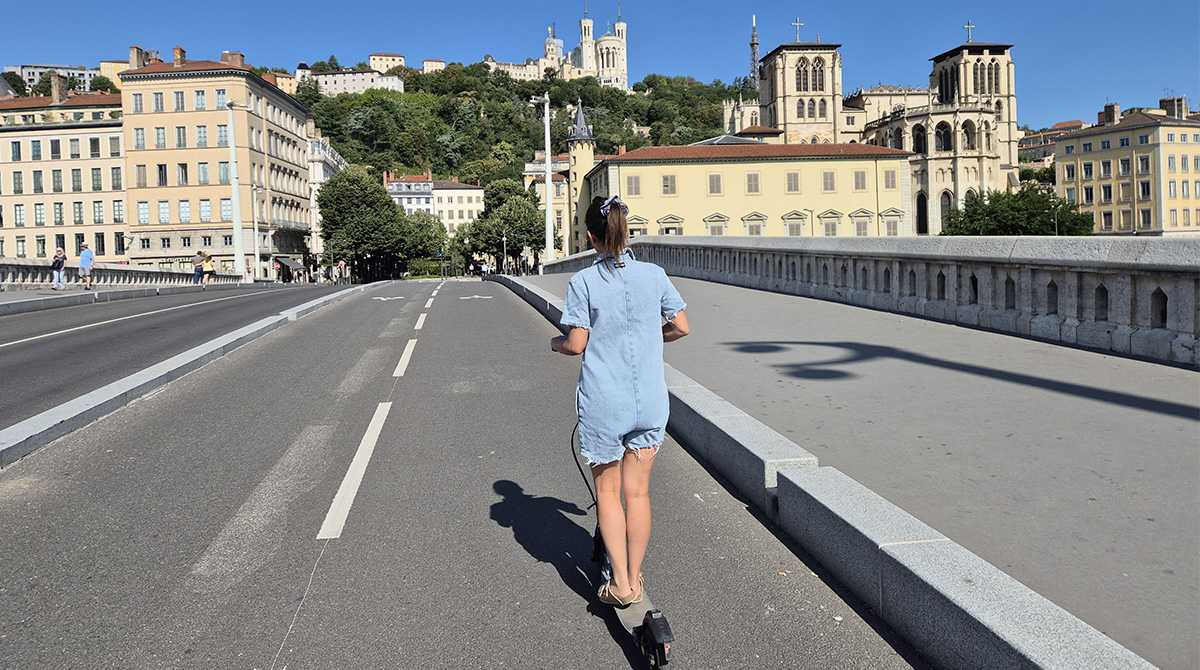 Jeune fille en trottinette électrique sans casque sur une piste cyclabe avec bâtiments et Fourvière en arrière plan