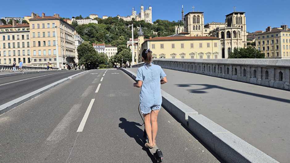 jeune fille sur une trottinette électrique sans casque de dos sur une piste cyclabe avec bâtiments et Fourvière en arrière plan